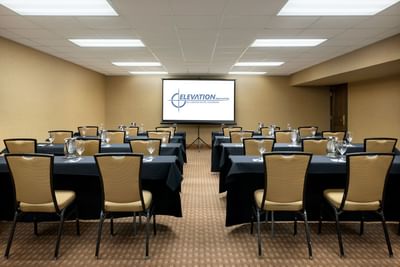 A conference room setup with multiple tables and chairs facing a large screen at Elevation Hotel and Spa.