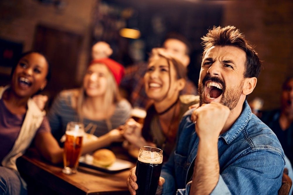 A group of people seated around a table holding pints of beer cheer at something out of frame.