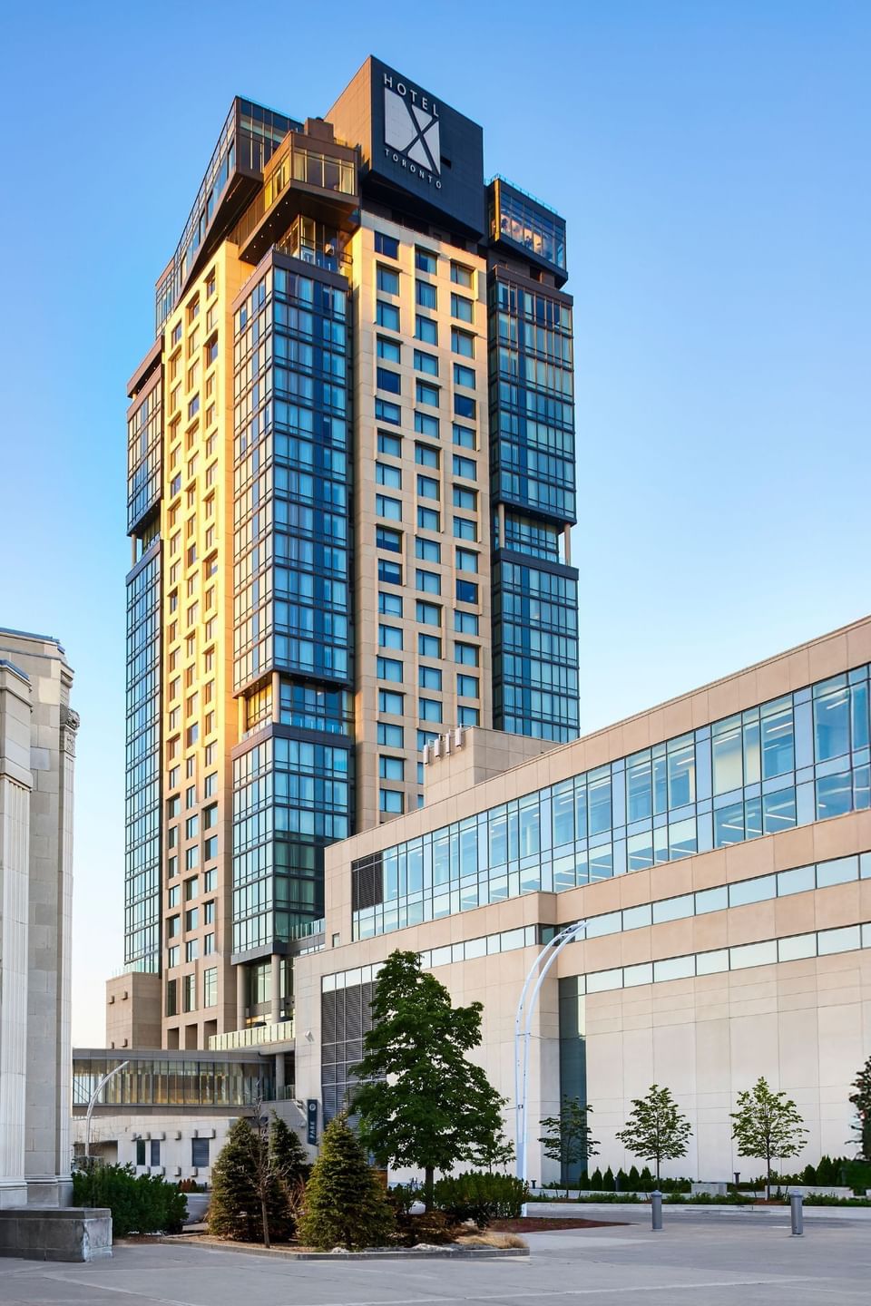 Tall modern glass hotel tower facing clear blue sky with trees and paved entrance walkway at Hotel X Toronto venue