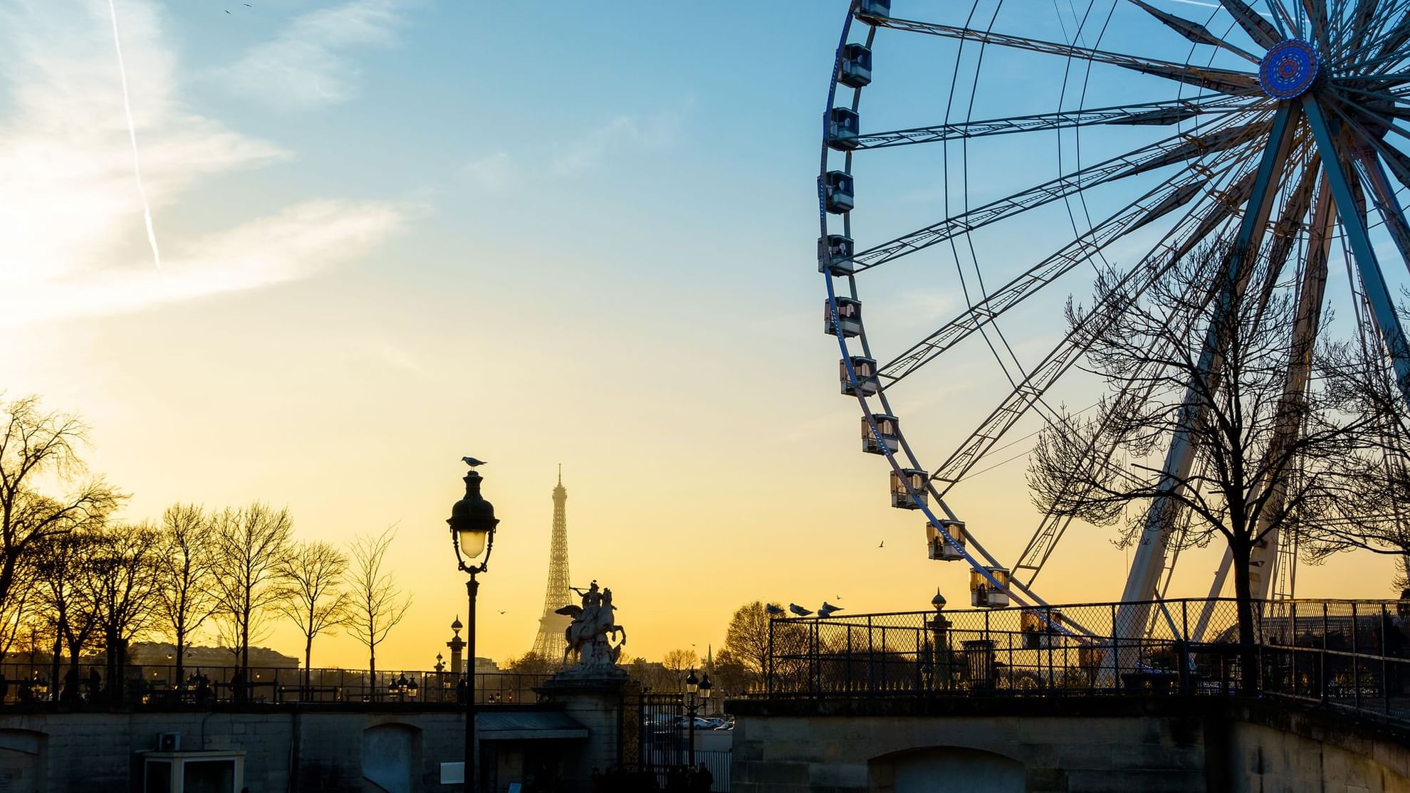 Sunset view of Paris featuring the Eiffel Tower, a Ferris wheel, and silhouetted trees near Warwick Hotels and Resorts