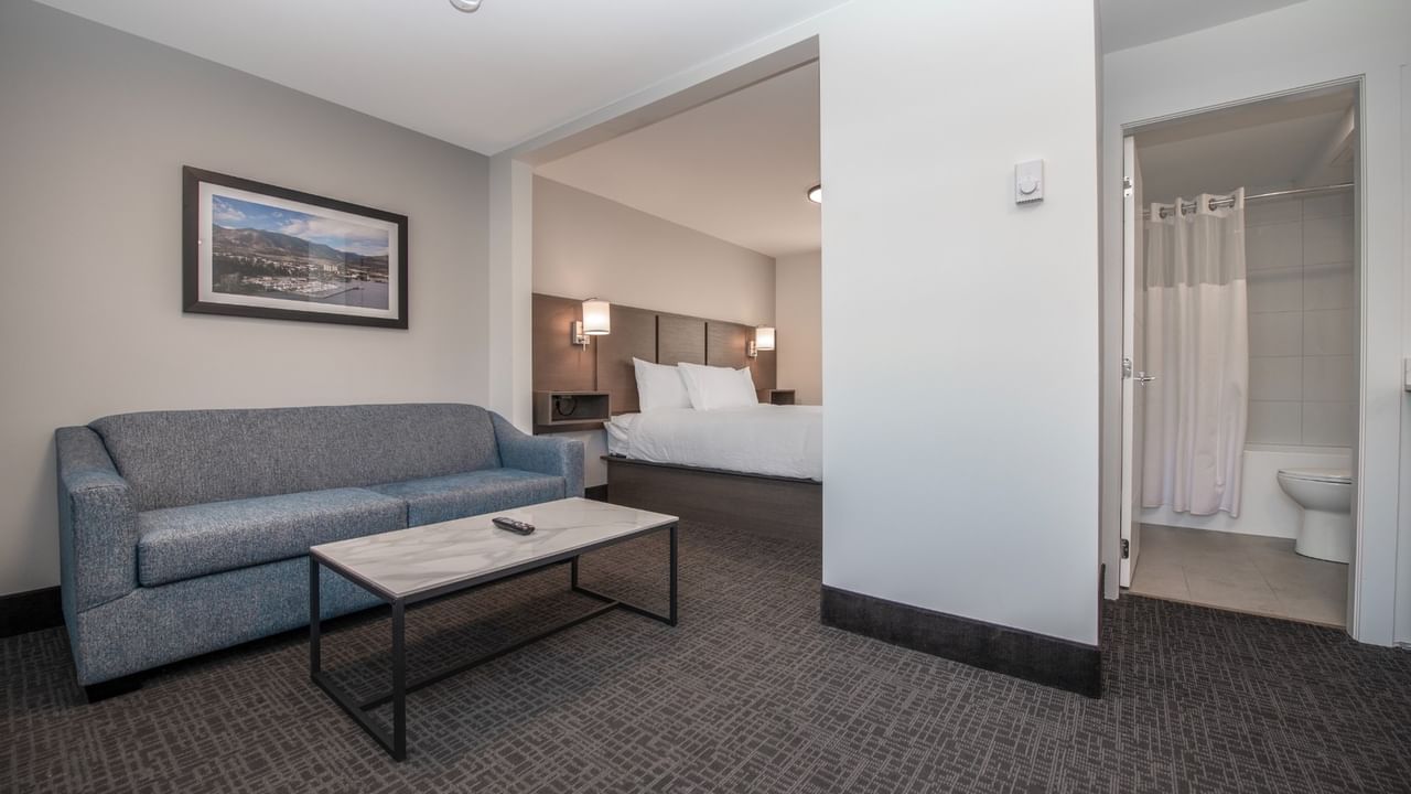 Hotel suite living room with couch, coffee table, and a bedroom with a bed and bathroom.