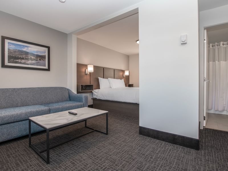 Hotel suite living room with couch, coffee table, and a bedroom with a bed and bathroom.