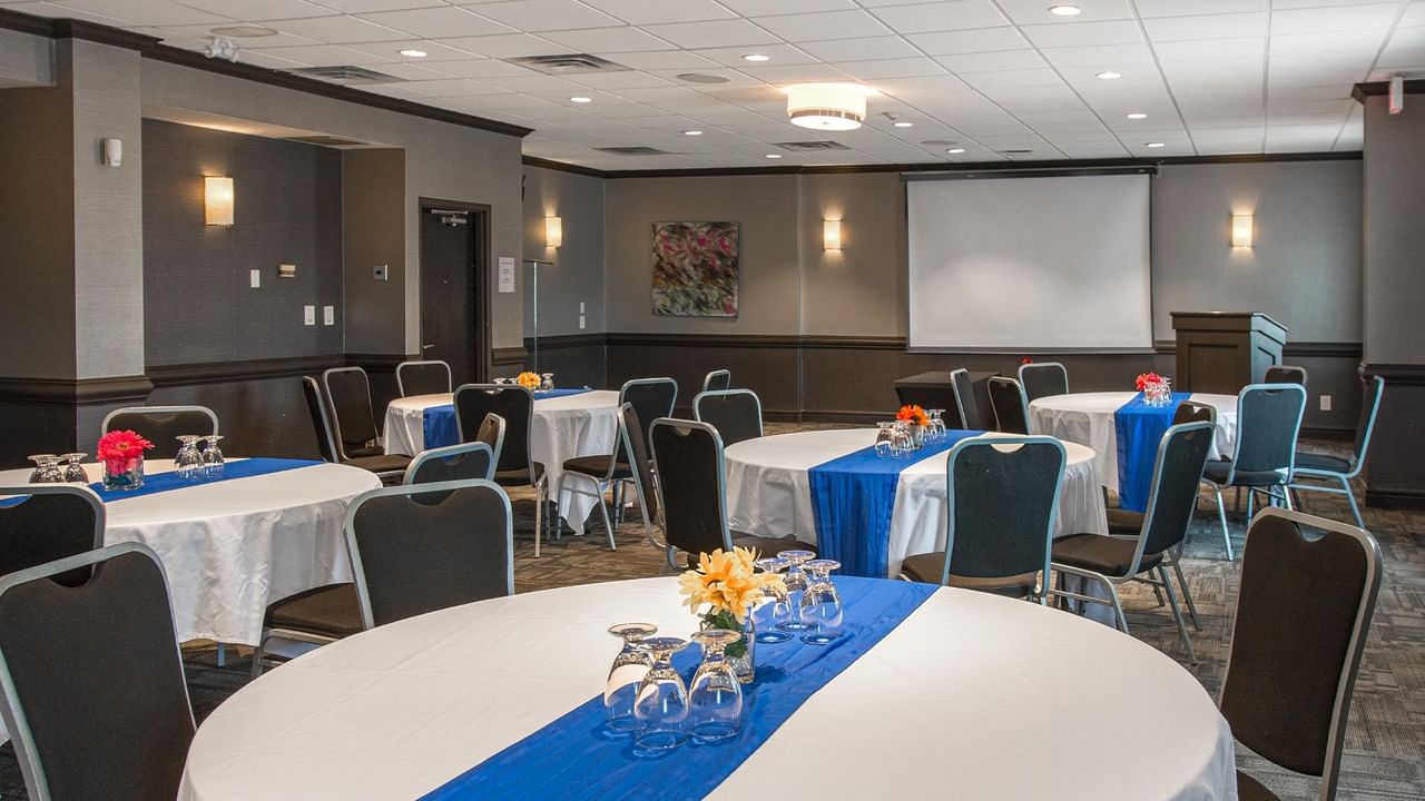 Meeting room with round tables, blue runners, floral centerpieces, and projection screen at Coast Calgary University Area Hotel by APA
