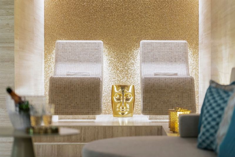 Two white chairs with gold face statue between them in a room with champagne and blue pillows.