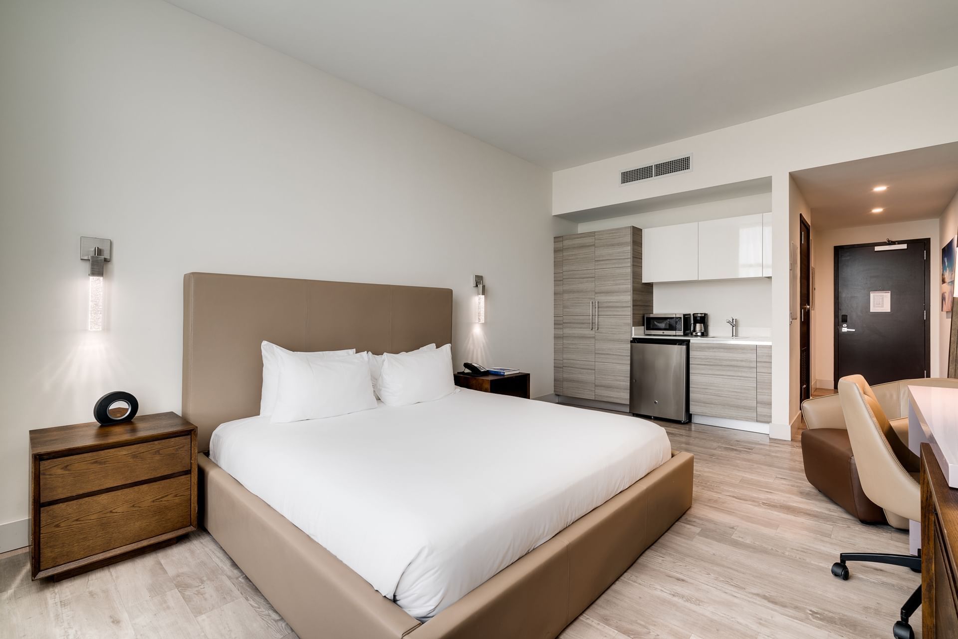 A modern hotel room with a king-sized bed, kitchenette, and desk.