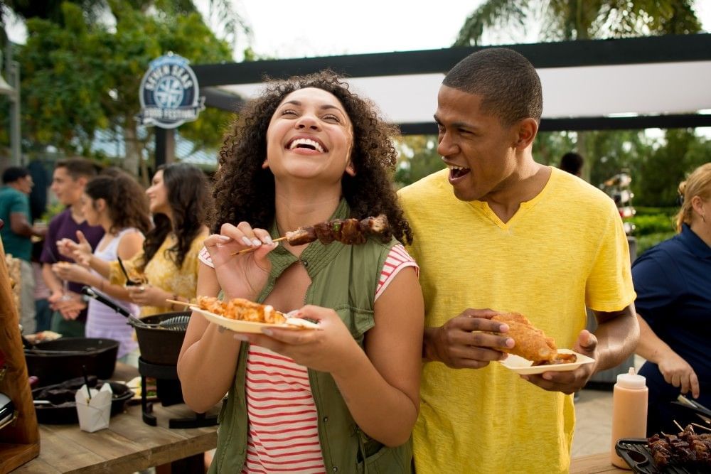 A couple smiling and laughing while holding plates of food outdoors. 