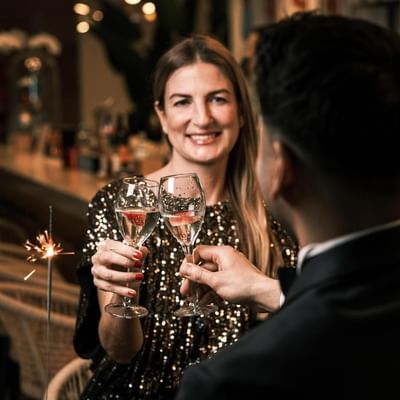 Frau und Mann in eleganter Kleidung stoßen mit Champagnergläsern an, umgeben von Glitzer und Lichtern.