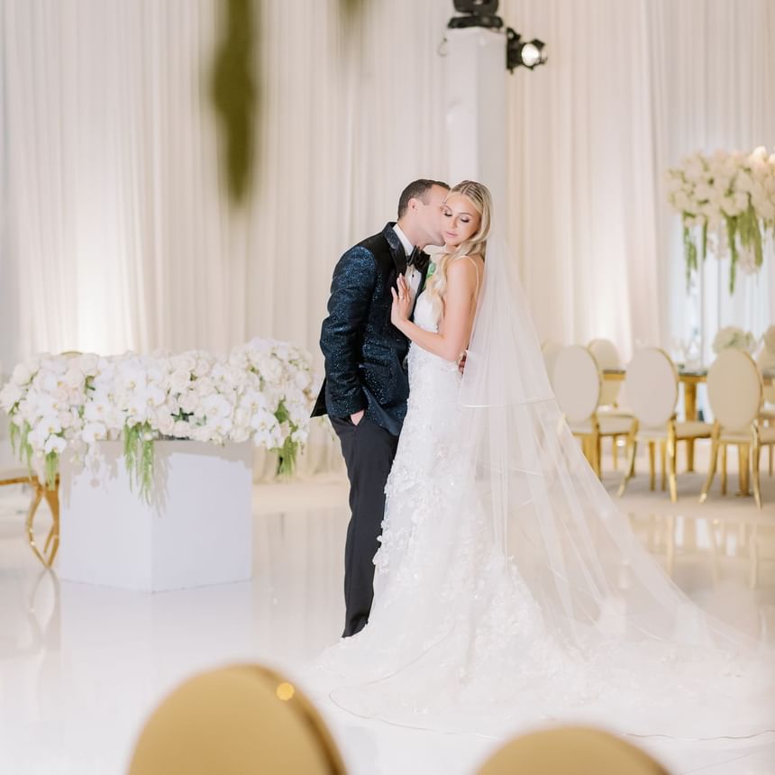 A wedding couple hugging & kissing in a ballroom at Luxe Sunset Boulevard Hotel