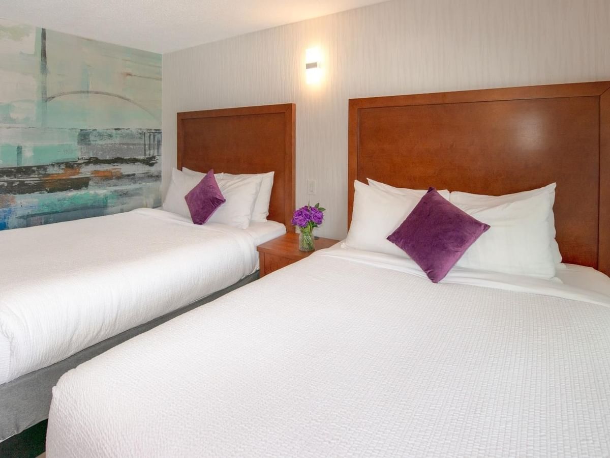 Two queen beds with white linens and purple pillows, wooden headboards and a nightstand between them.