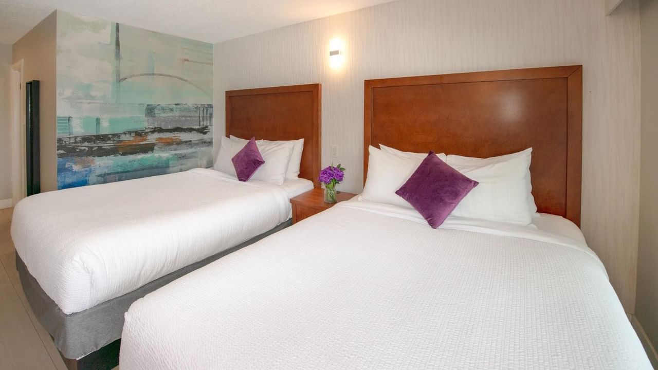 Two queen beds with white linens and purple pillows, a vase with flowers and a mural on the wall.