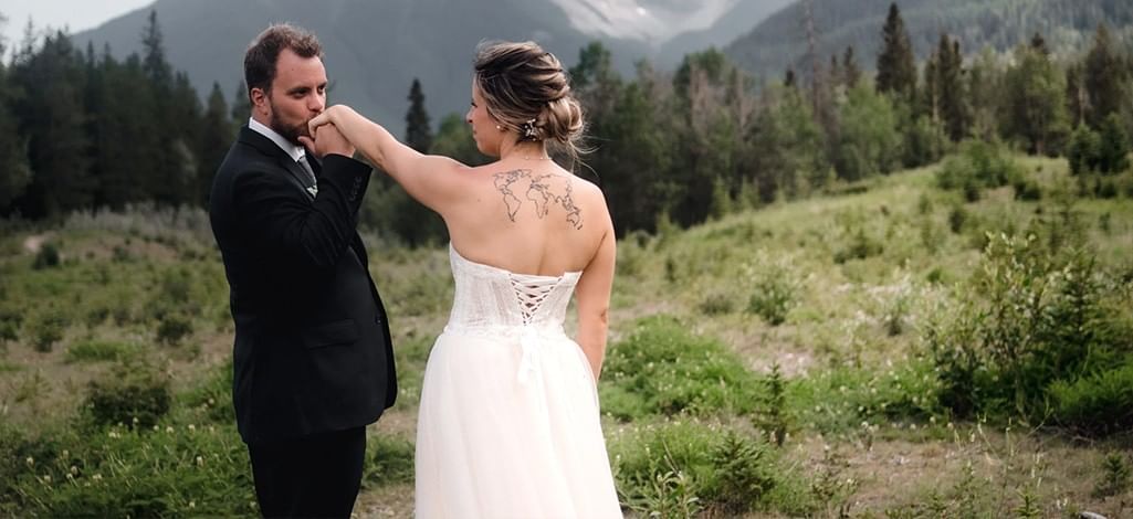 A groom kisses a bride's hand in front of the mountains after their wedding in Canmore.