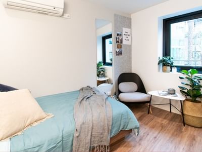 A modern single bedroom with a bed, chair, small table, large window, and two potted plants.