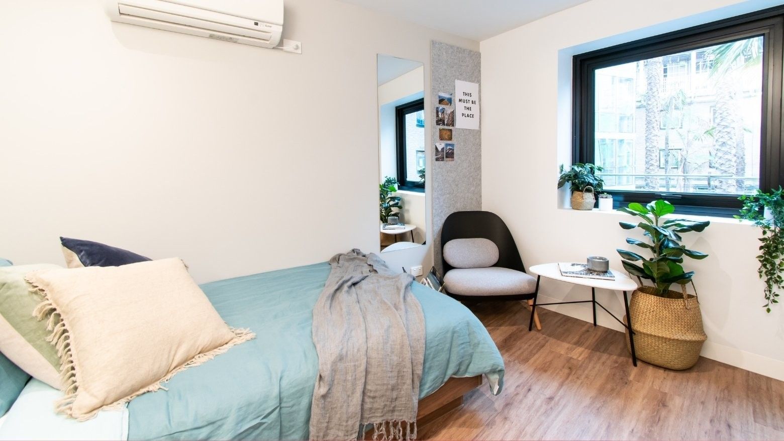 A modern single bedroom with a bed, chair, small table, large window, and two potted plants.