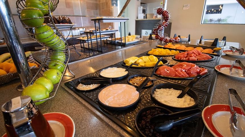 Buffet with yogurt bowls and granola beside sliced fruit and a green apple stand in Palmira Cafeteria at Araiza Calafia