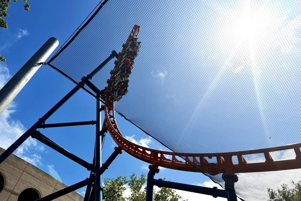 A roller coaster car of people hangs suspended at the end of a coaster track behind netting on a sunny day.