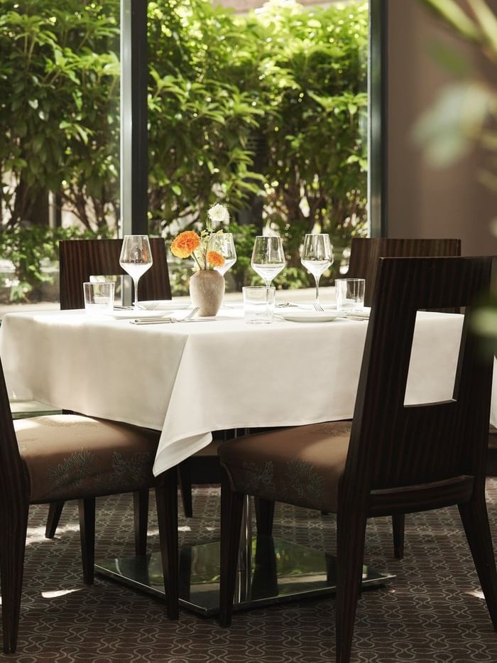 Elegant dining table with four chairs, white tablecloth, floral centerpiece, and large windows with garden view.