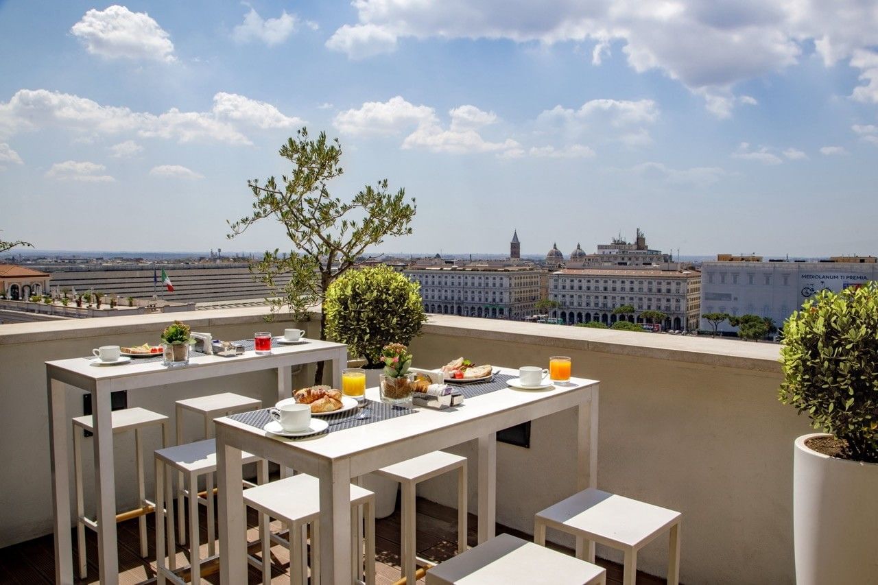 Breakfast buffet with fresh fruit, pastries, and juices in a modern rooftop lounge with outdoor seating and city skyline views.