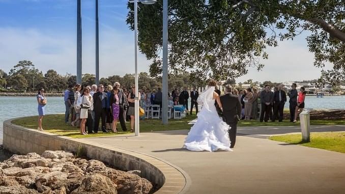 Bride and groom walk along an estuary path as wedding guests gather on a sunny day at The Sebel Mandurah