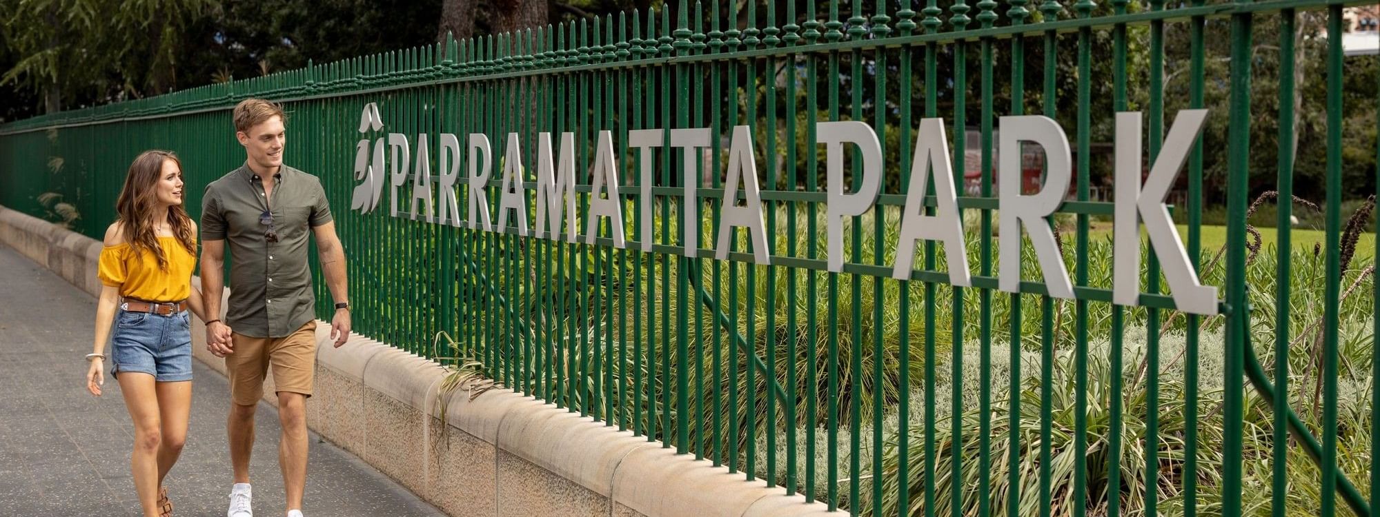 A couple walks along a path by a green fence labeled 
