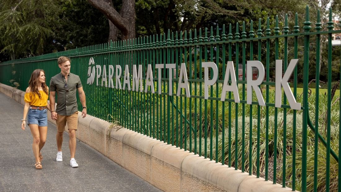 Couple walking hand-in-hand along a path next to a green metal fence labeled 