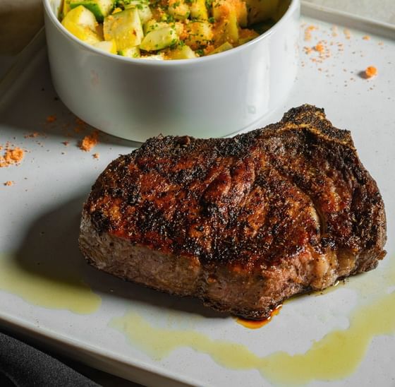 Seared steak on a white plate, served with a bowl of mixed diced vegetables and glass of beer at Cypress & Oak, Frankfort, KY