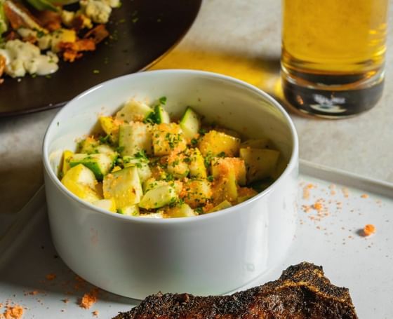 Seared steak on a white plate, served with a bowl of mixed diced vegetables and glass of beer at Cypress & Oak, Frankfort, KY