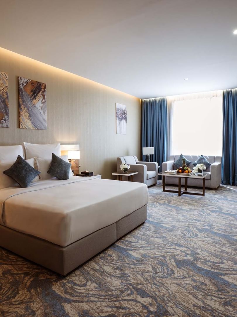 Large bed in Deluxe Room with paintings and a sofa under blue curtains by a window at Cantonal Hotel by Warwick Riyadh