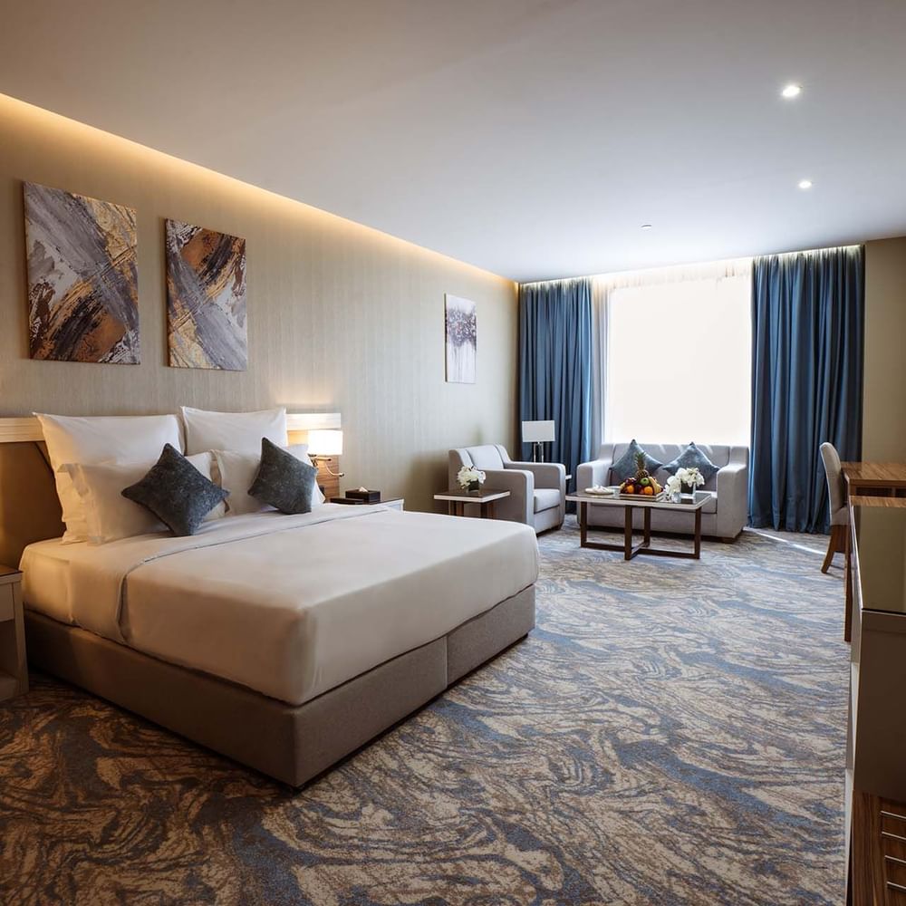 Large bed in Deluxe Room with paintings and a sofa under blue curtains by a window at Cantonal Hotel by Warwick Riyadh