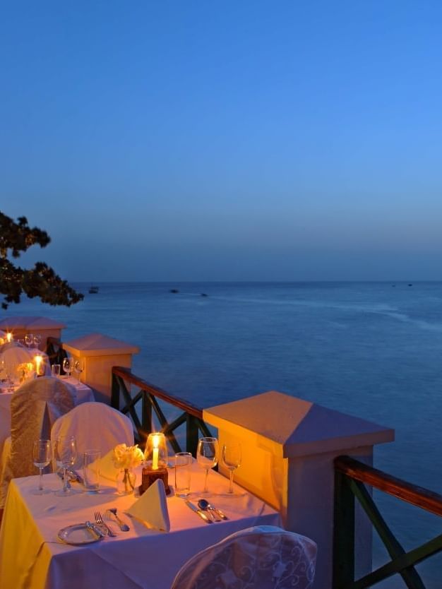 Candlelit dining tables arranged along the outdoor terrace balcony overlooking blue ocean at Serena Hotels & Resorts