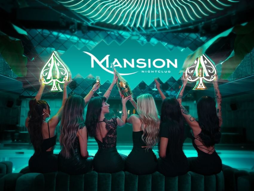Six women in black dresses raising champagne glasses in front of a Mansion Nightclub logo at Paradox Vancouver