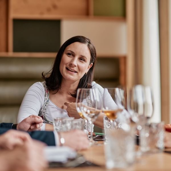 Eine Frau mit kurzem Haar sitzt an einem Tisch mit mehreren Gläsern und Wein, lächelt und unterhält sich.