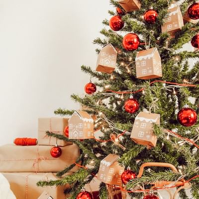 Ein geschmückter Weihnachtsbaum mit roten Kugeln, hausförmigen Anhängern und Geschenken am Boden.