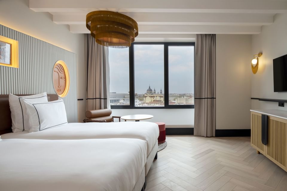 Beds under a golden oval light fixture by a round table with an armchair near a window in La Generous at Urban Hive Madrid