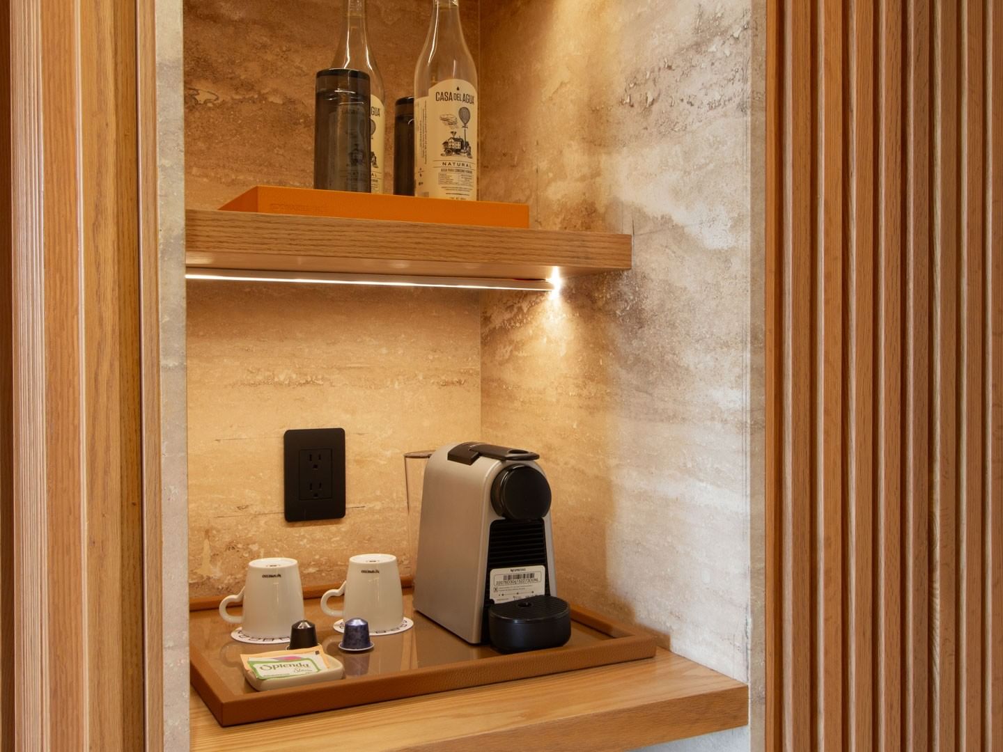 Estantería con cafetera, tazas y botellas en un espacio con paneles de madera.