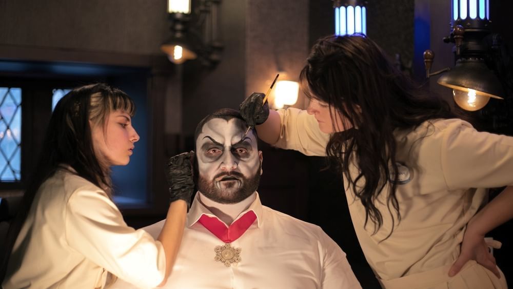 Two women in lab coats apply vampire makeup to a man staring into the distance.