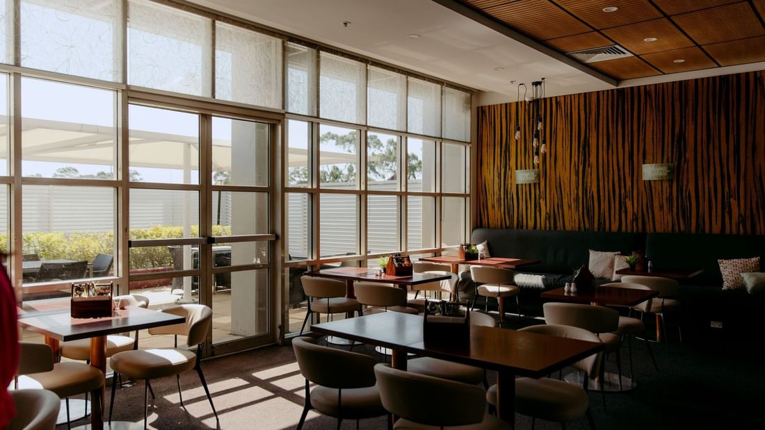Empty restaurant with tables, chairs, and large windows, overlooking an outdoor patio and greenery.