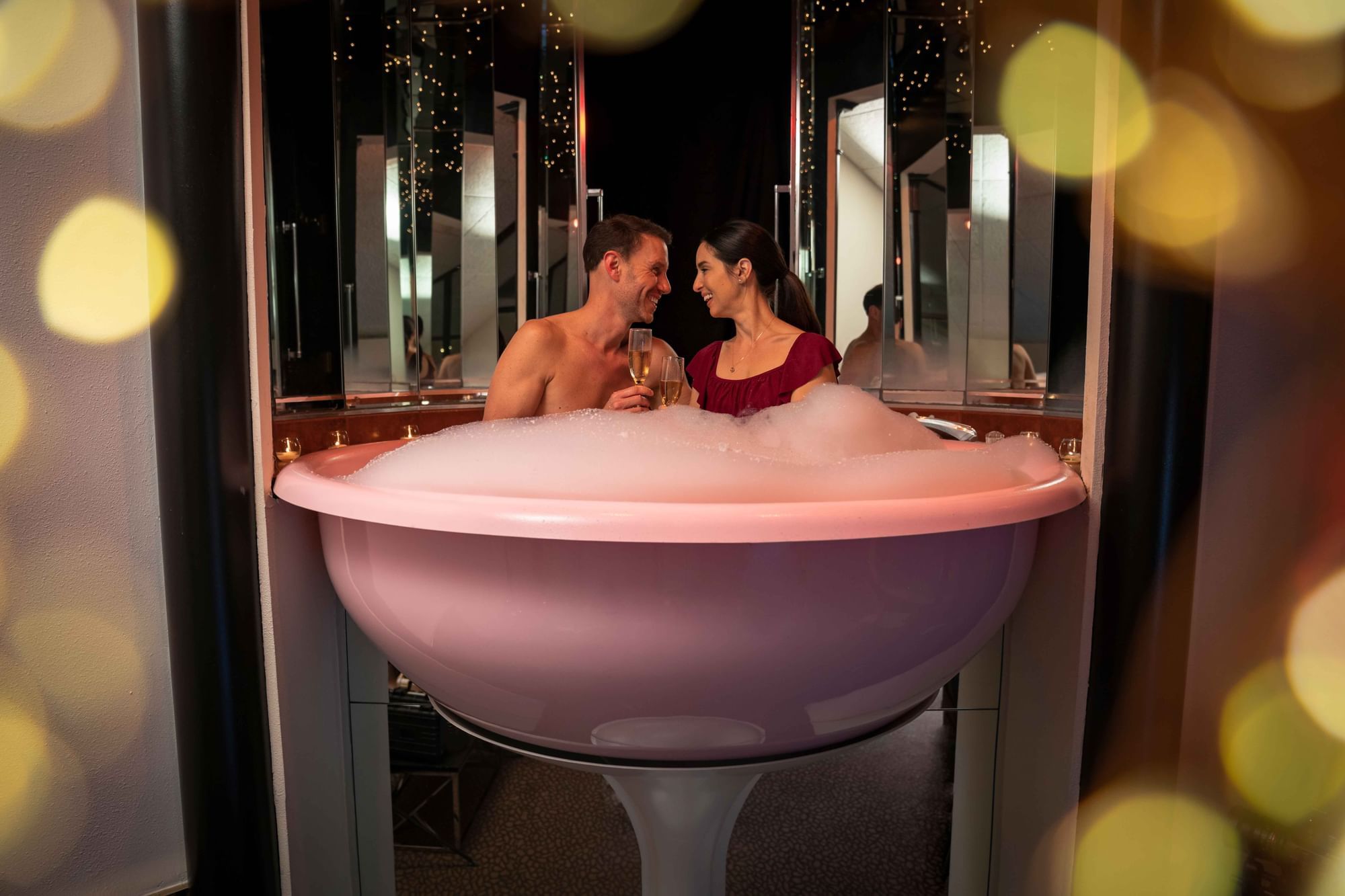 A couple enjoys a romantic bubble bath, holding drinks, surrounded by shimmering lights at Cove Pocono Resorts
