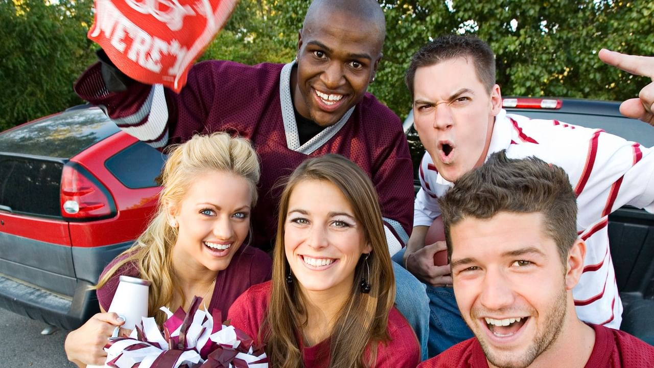 Five happy people in team jerseys, one holding a pompom, posing by a red car.