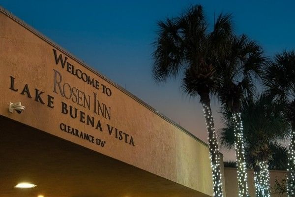 An exterior sign for Rosen Inn Lake Buena Vista beside palm trees wrapped in white Christmas lights. 