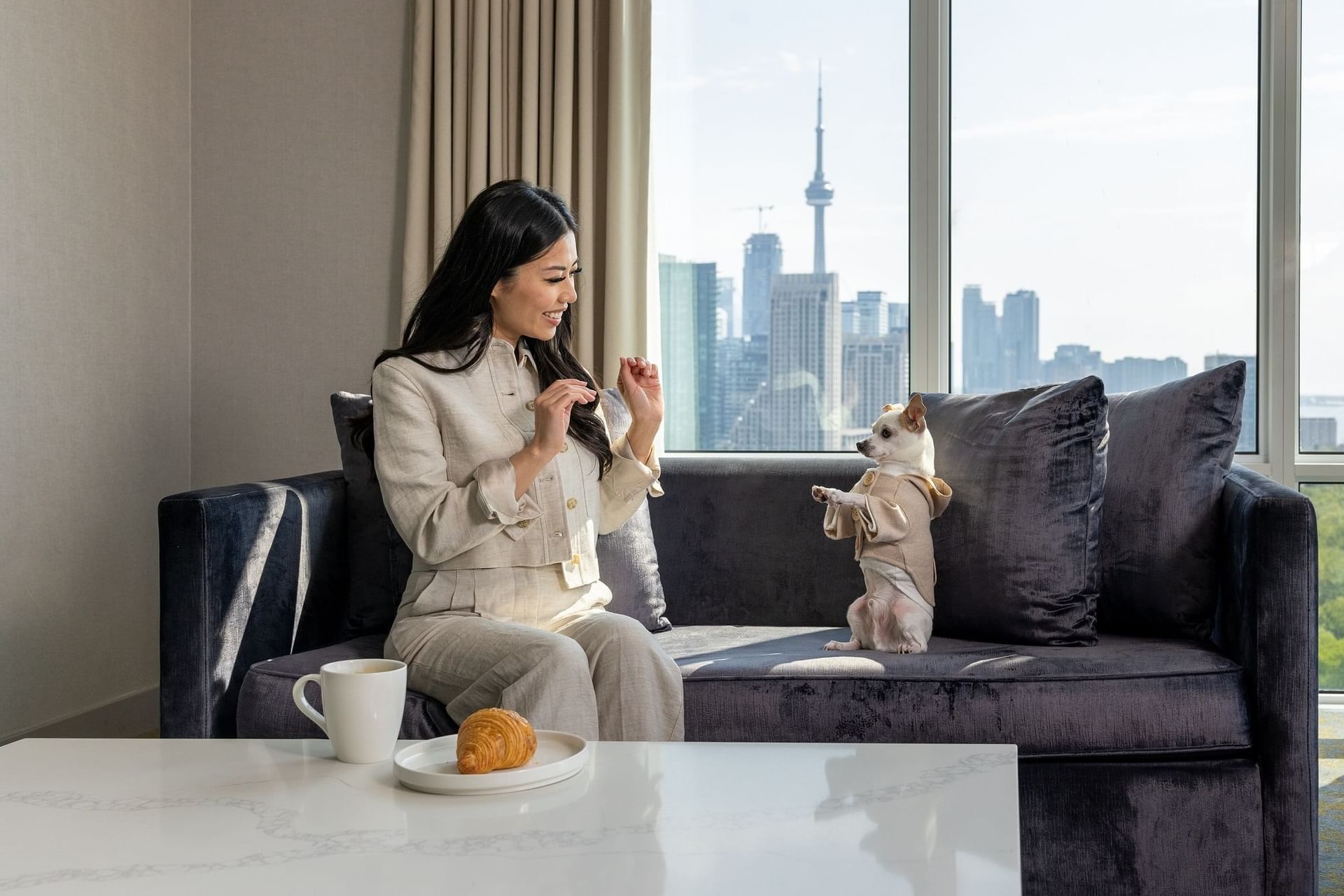 Woman sitting on sofa playing with a dog with coffee and croissant on a table facing CN Tower skyline at Hotel X Toronto