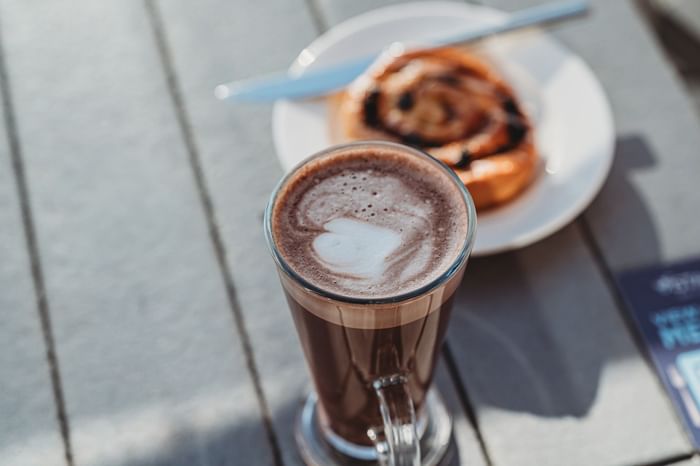 Cup of hot chocolate with heart-shaped foam on a table next to a plate with a pastry at hotel Vista Bar Cafe