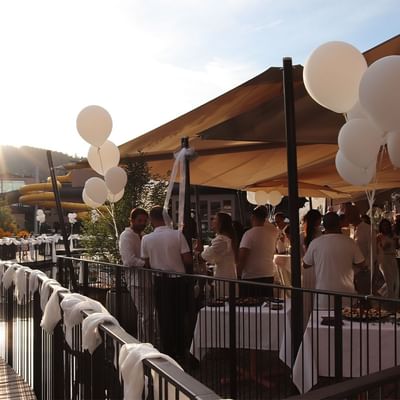 Outdoor event with white balloons, canopy, tables, and guests in white attire under a sunny sky.