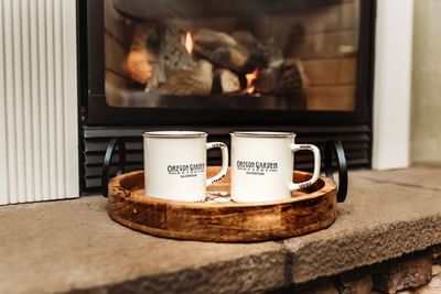 Two Oregon Garden Resort mugs on a wooden tray in front of a fireplace with burning wood.