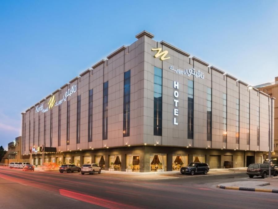 Modern hotel building with illuminated signage and parked cars in front at dusk.