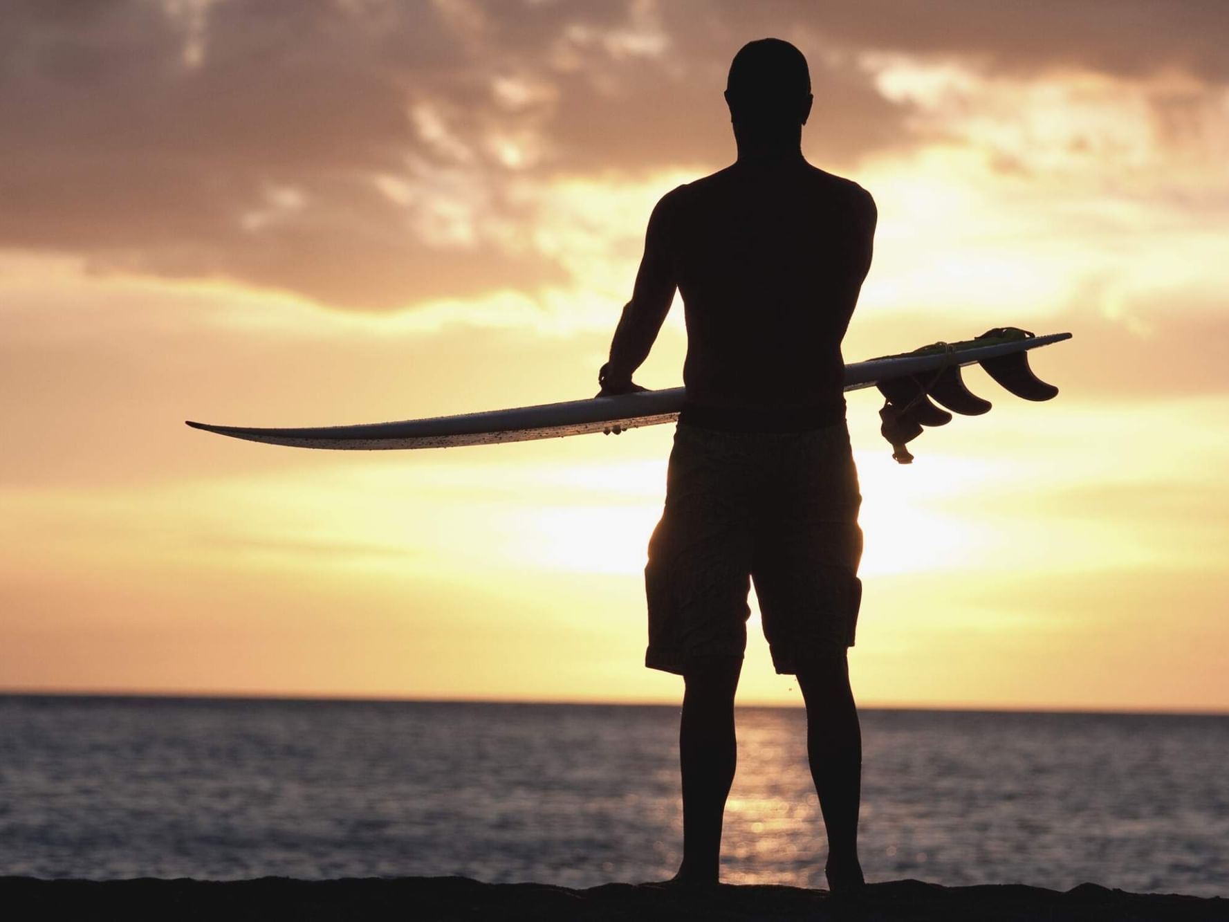 Man silhouette holding surfboard at sunset on the beach for Best Time to Visit Oahu.