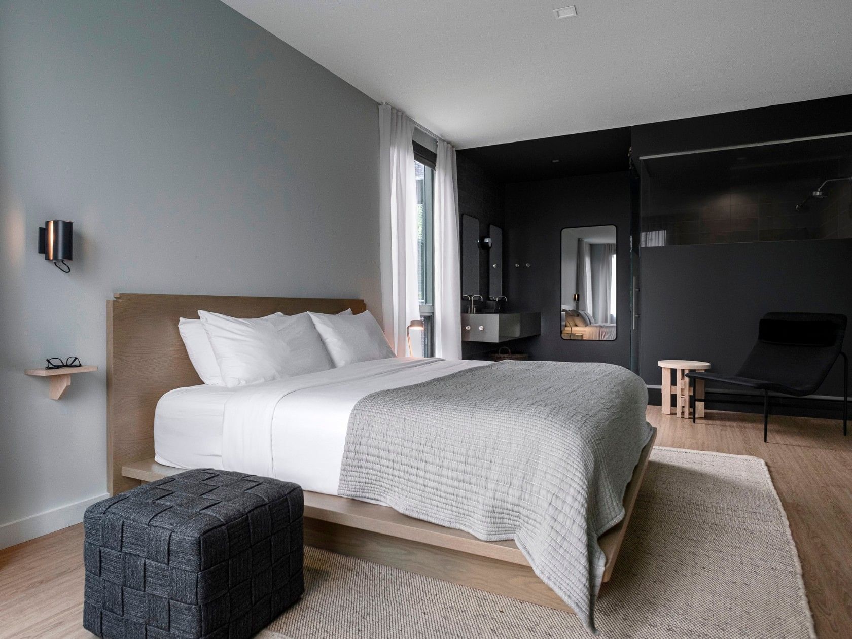 Bedroom with a bed, gray blanket, four white pillows, black ottoman, and black chair near a bathroom.