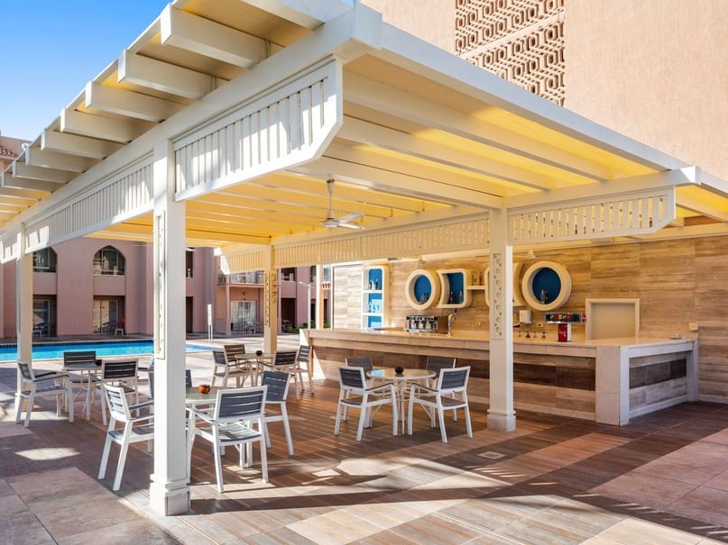 Outdoor dining area with tables, chairs, and a bar under a white pergola near a swimming pool.