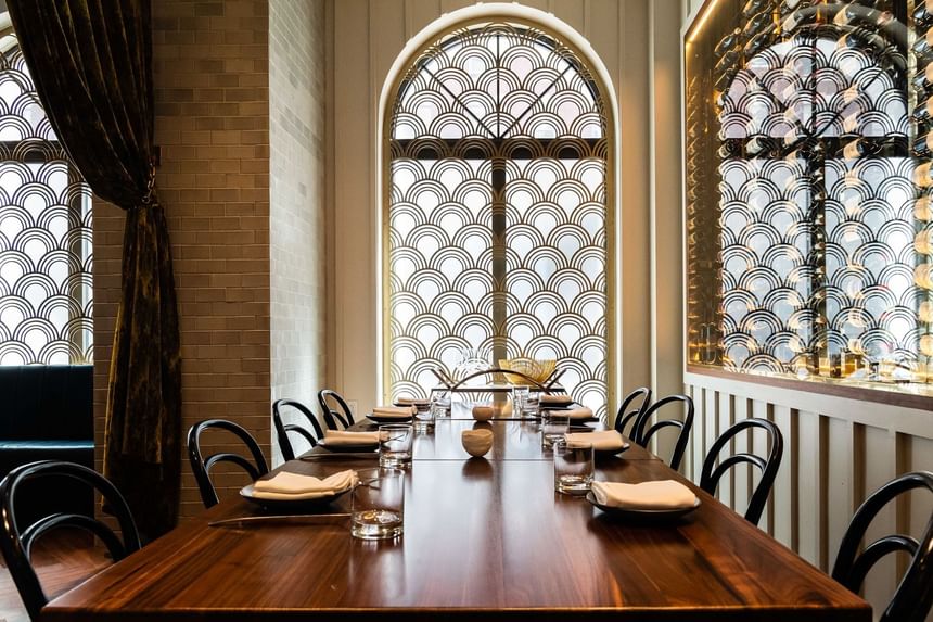 Long wooden dining table set with chairs beside arched patterned windows and a glass wine display wall at Hotel Bijou