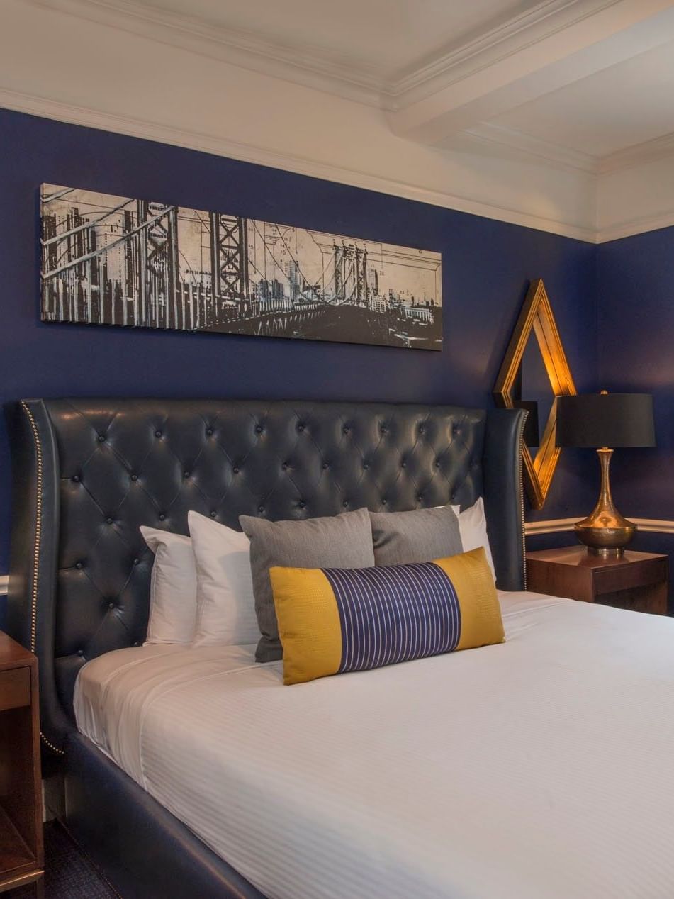 Bold blue bedroom with a leather tufted headboard & gold mirrors, and city bridge art in Randolph Suite at Warwick New York