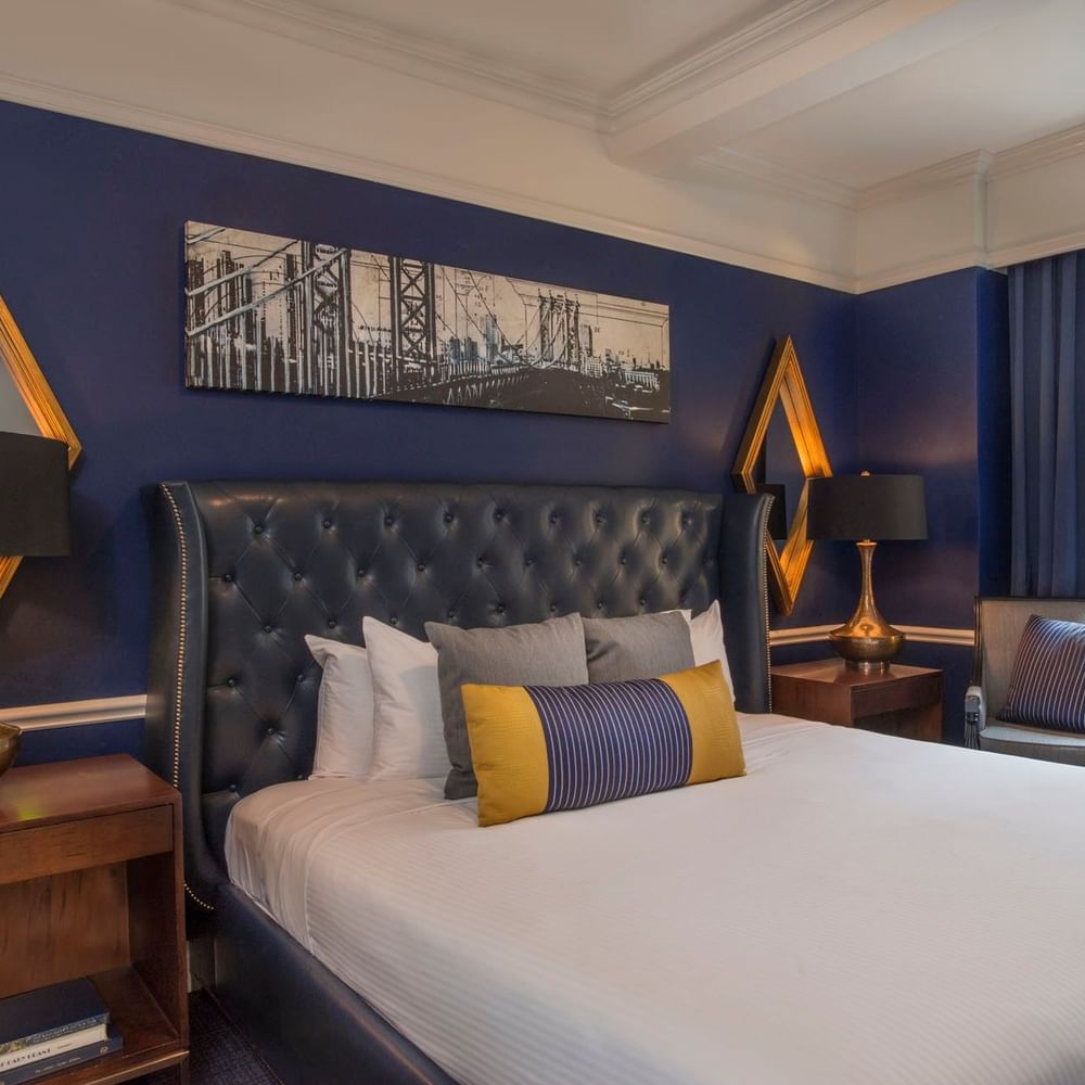 Bold blue bedroom with a leather tufted headboard & gold mirrors, and city bridge art in Randolph Suite at Warwick New York